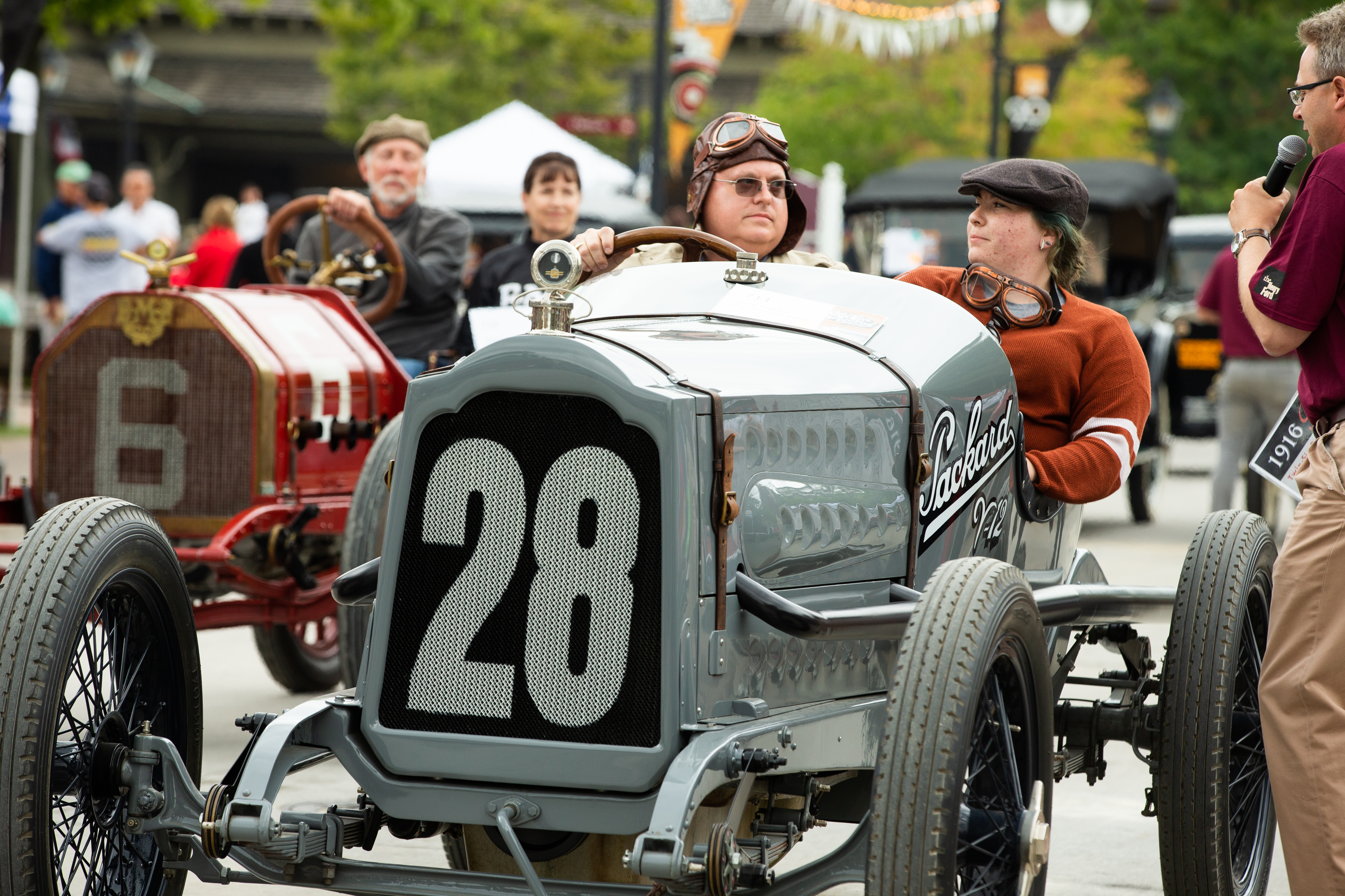 Two people in vintage car in foreground and another two in another car in background; additional cars in the distance