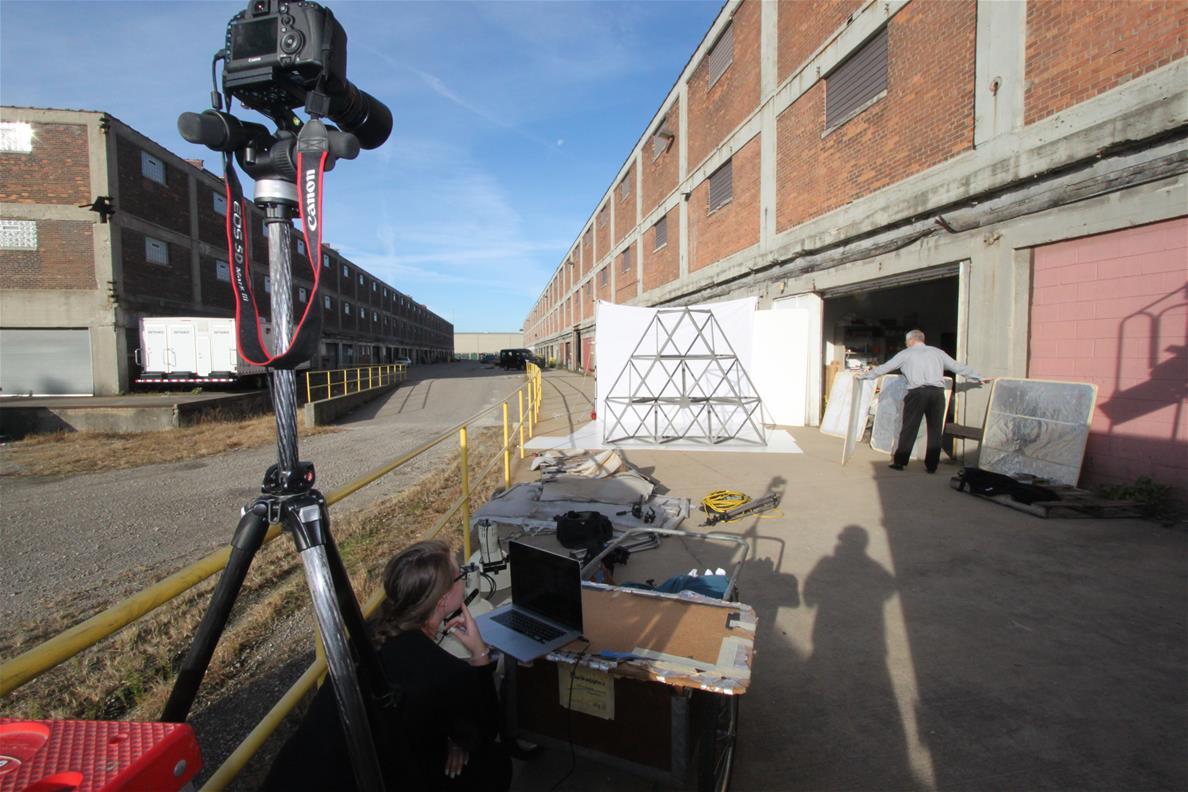 Photography staff and volunteers shooting the geodesic truss on location at a storage building Camera on tripod in foreground pointing toward white paper with metal truss in front of it alongside a large red brick building