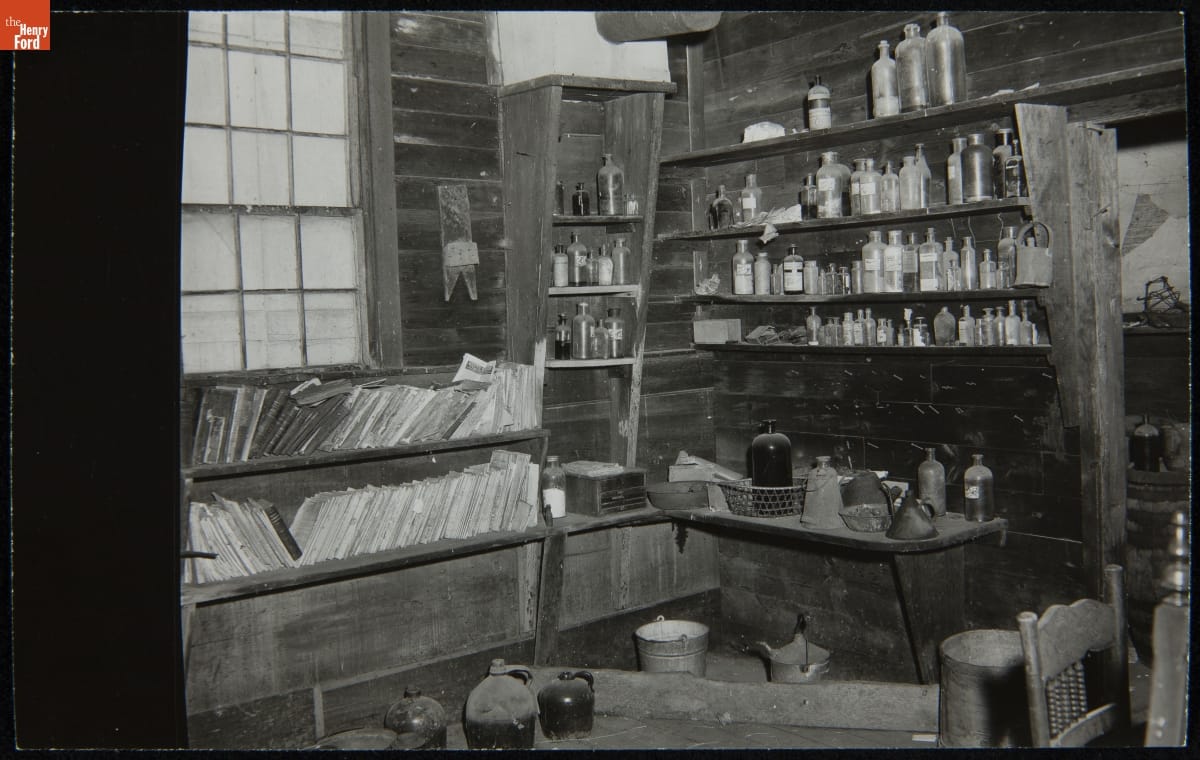 Room interior containing shelves and tables covered in books, bottles, jugs, and other items