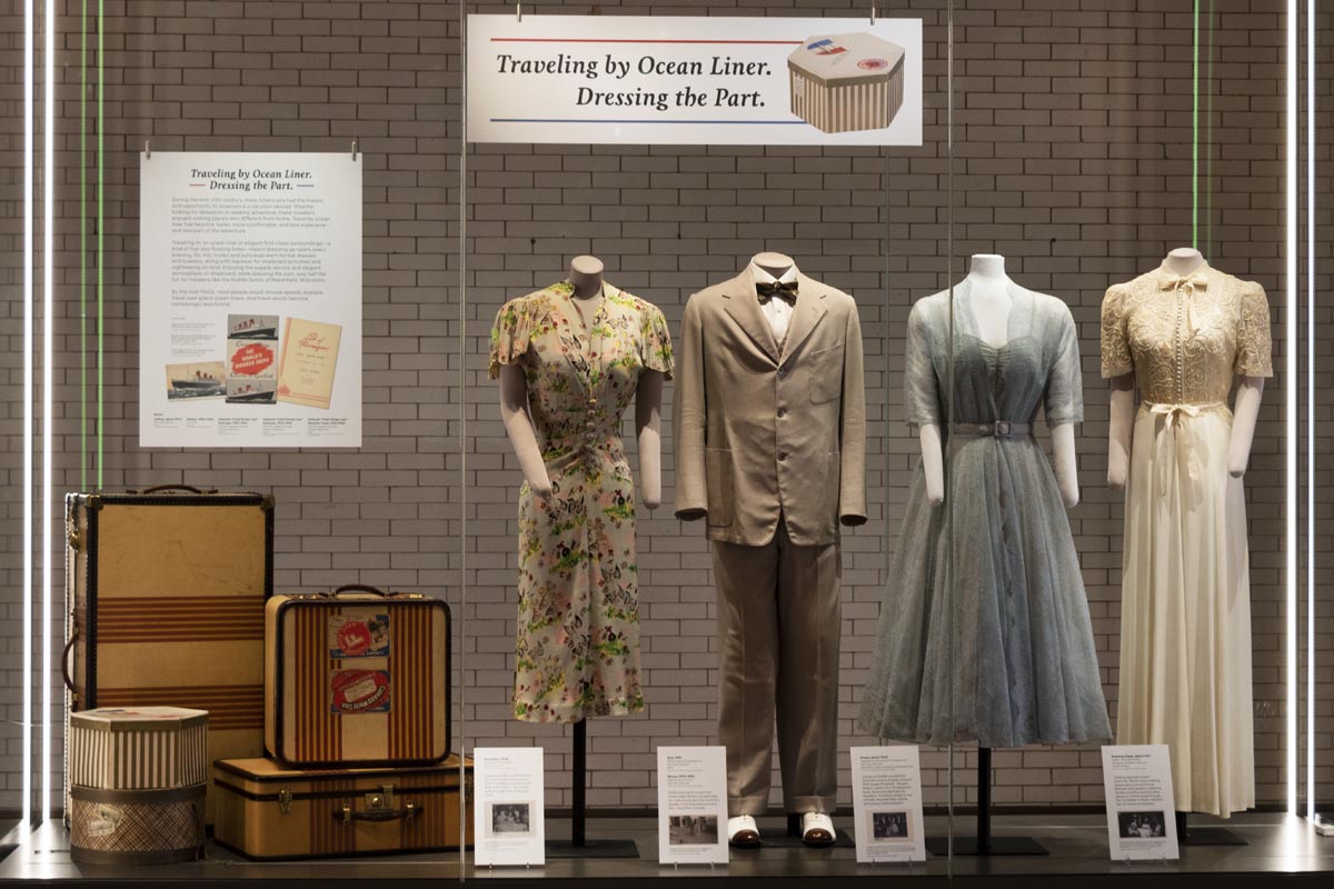 What We Wore: Traveling by Ocean Liner Large museum display case containing four mannequins wearing clothing, luggage, and signage