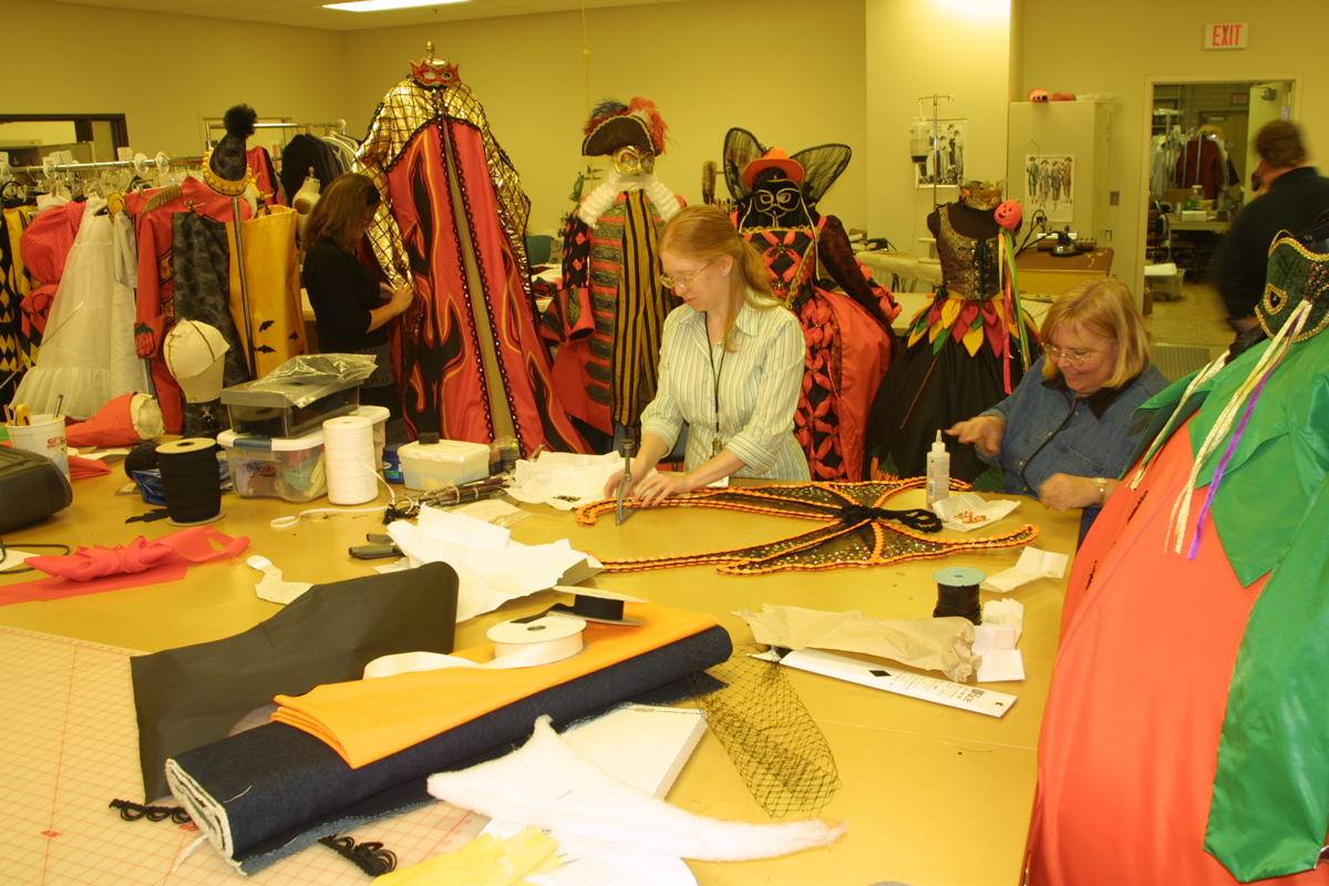 Two women work at a large table filled with fabric bolts and sewing notions; elaborate costumes hang in the background