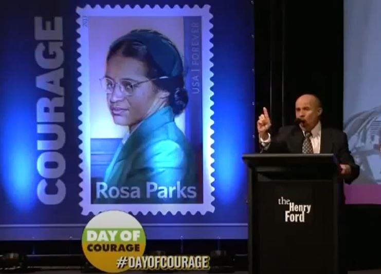 Man in suit stands at podium with hand pointed at the audience in front of backdrop of large Rosa Parks stamp