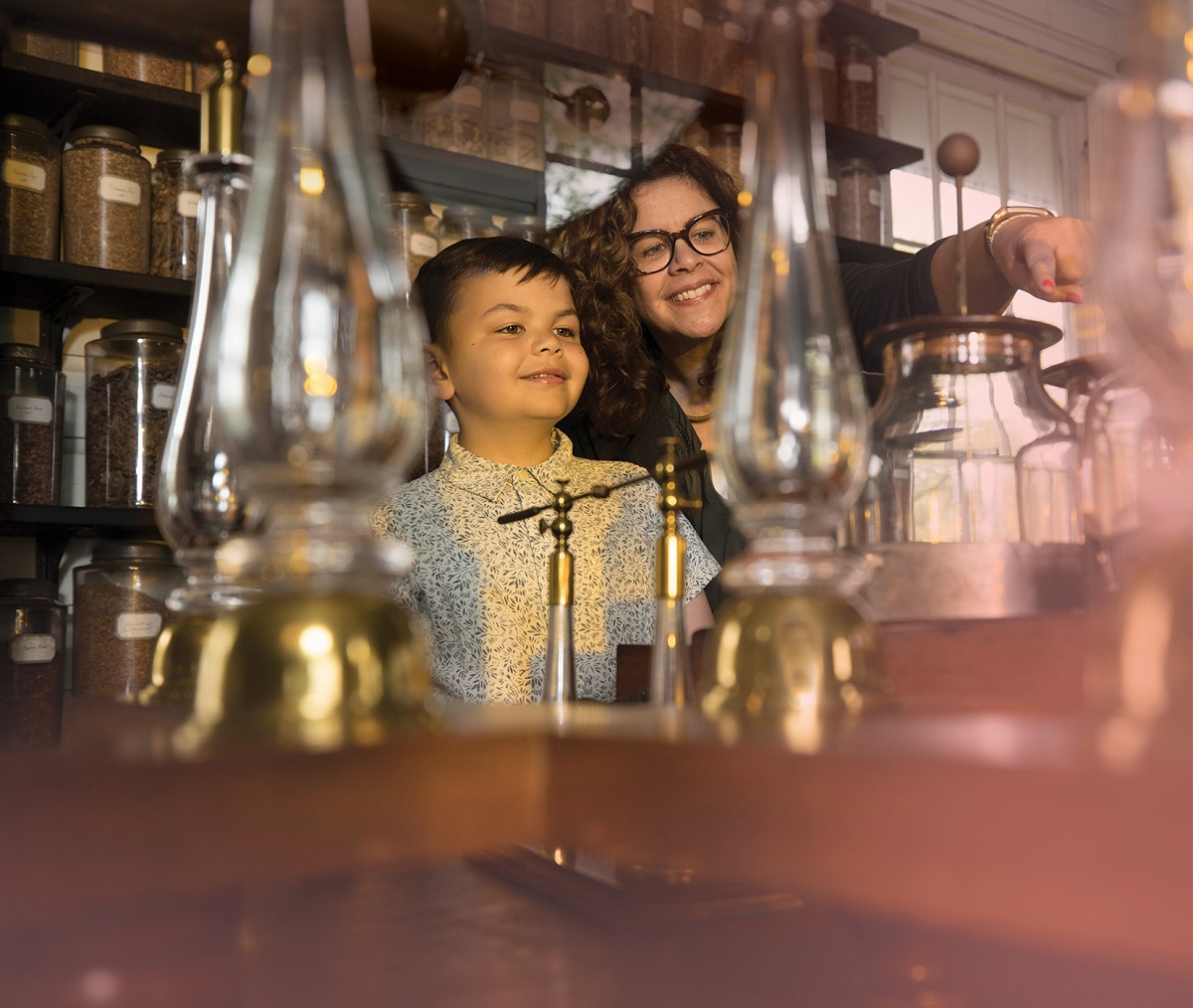 Mary Aviles and her son, Mati A woman and young boy smile, point, and look at a table filled with glass jars and lamps, with shelves of more glass jars behind them