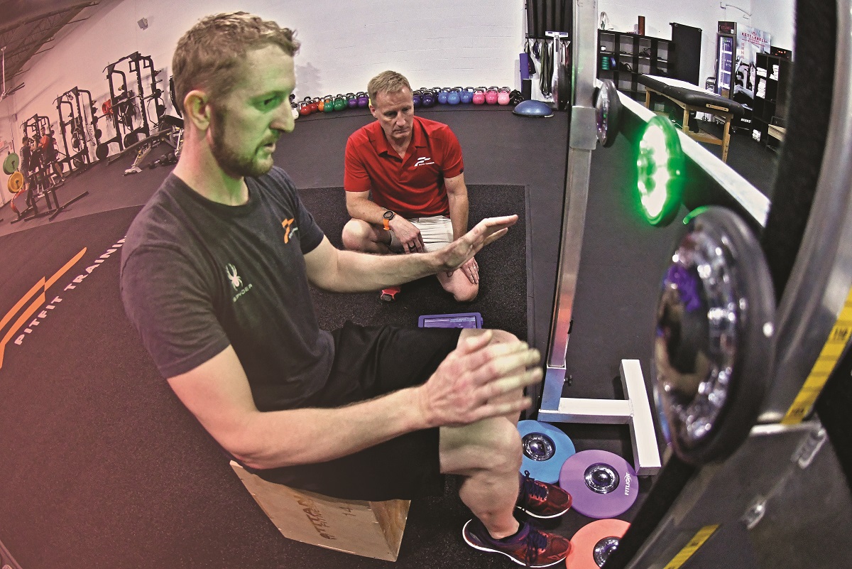 Driver Charlie Kimball training at PitFit Man in t-shirt and shorts sits in front of lighted device in gym with hands raised as another man kneels next to him