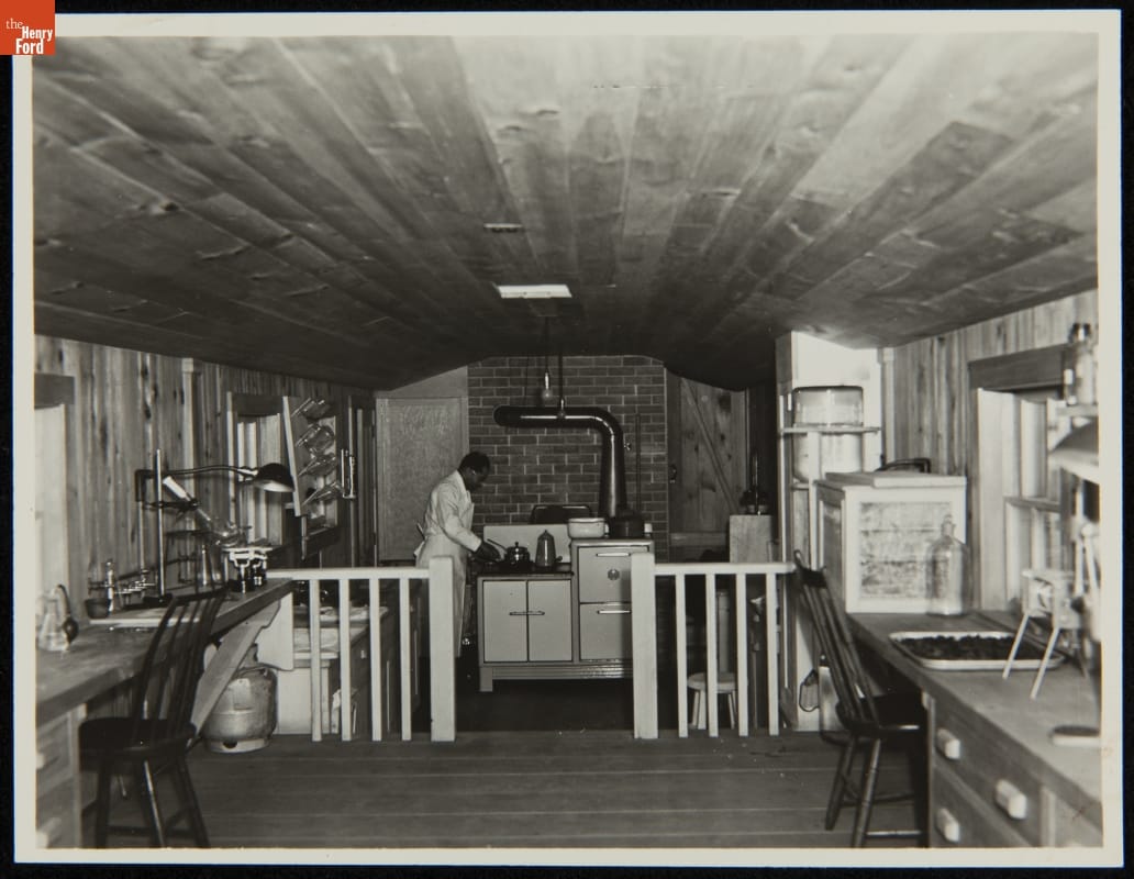 Man in an apron works at a stove at the far end of a room filled with workspaces and equipment