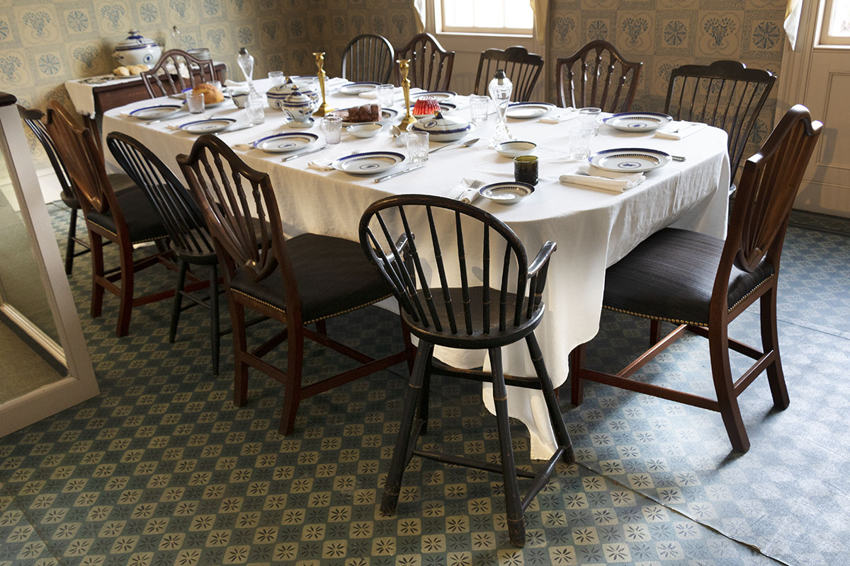 Room with patterned floor and walls containing a large, set table with many mismatched chairs