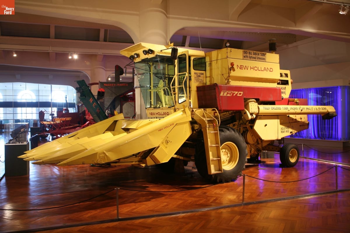 New Holland TR70 Axial Flow Combine, 1975 Large yellow piece of agricultural equipment with large tines at front below a cab, in a large space with wooden floor and other agricultural equipment
