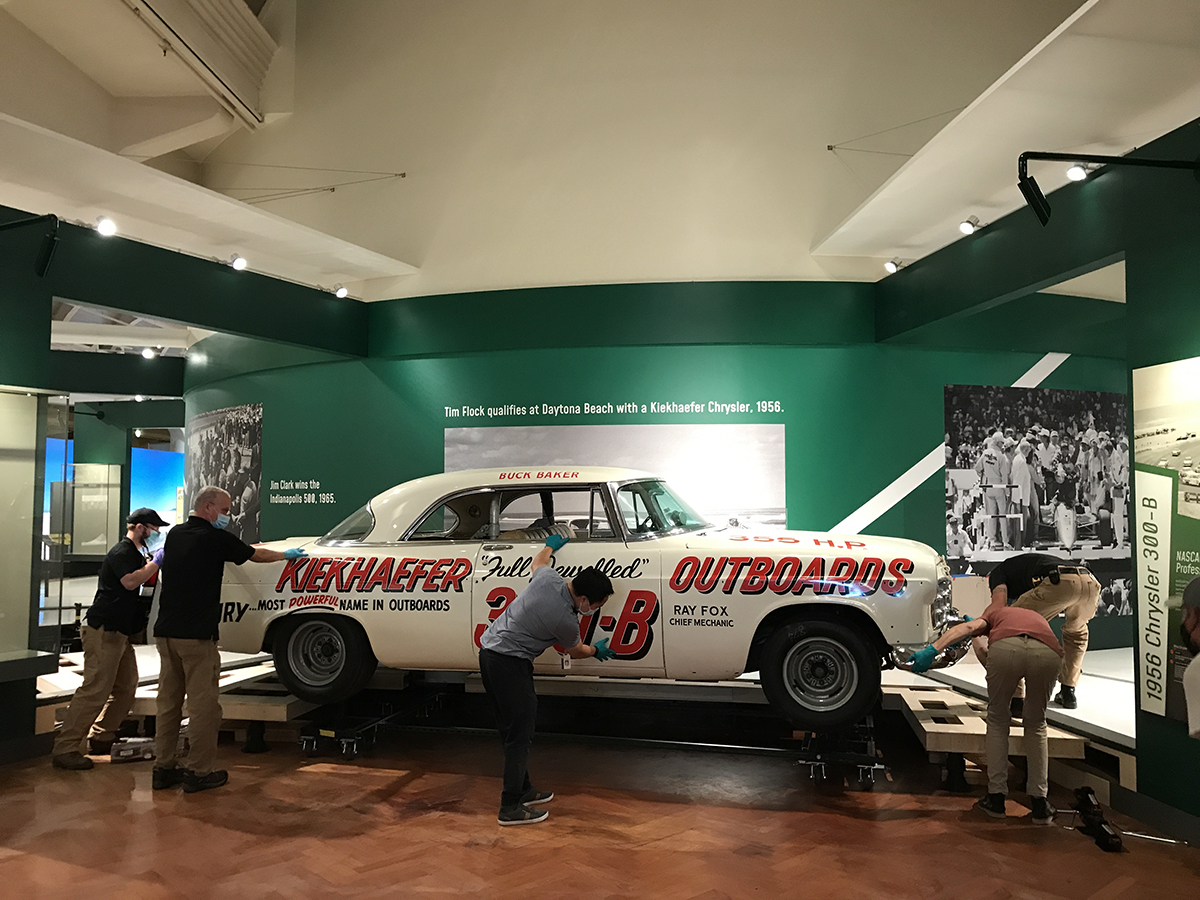 1956 Chrysler 300B Stock Car rolling into its display position. White car with bold red text along the side with several people guiding it into place in an exhibit