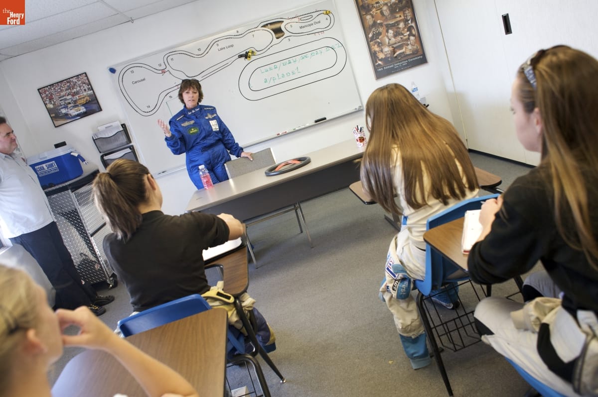 Lyn St. James at her Complete Driver Academy, 2008 Woman in blue jumpsuit stands behind a table, in front of a whiteboard, at the front of a room with young women sitting in school desks