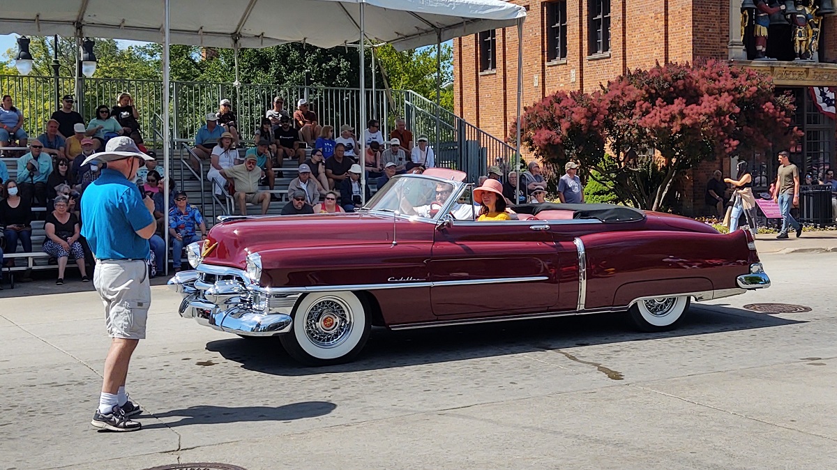 A beautiful 1953 Cadillac Series 62 convertible at Motor Muster 2022 A long, burgundy-colored convertible with dramatic lines drives past a man with a microphone in front of grandstands with seated spectators