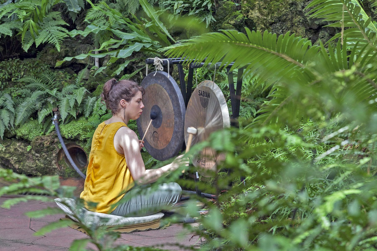 Lavender Suarez Woman in yellow top sits on cushion and hits two hanging gongs with mallets on a stone patio surrounded by foliage