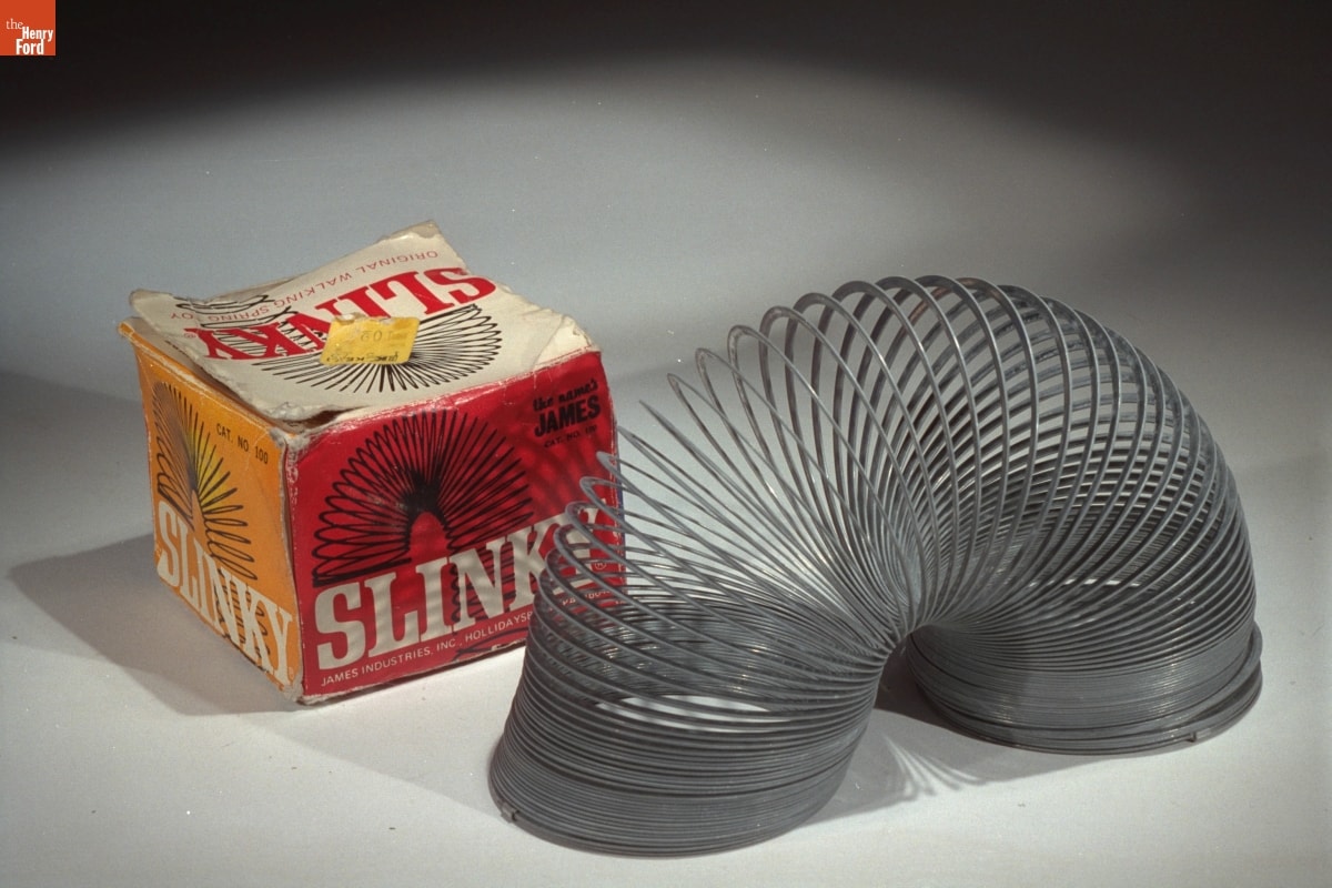 "Slinky," 1970-1980 Image with metal coil (Slinky toy) in arch shape in front of yellow and red with text and image of Slinky coil