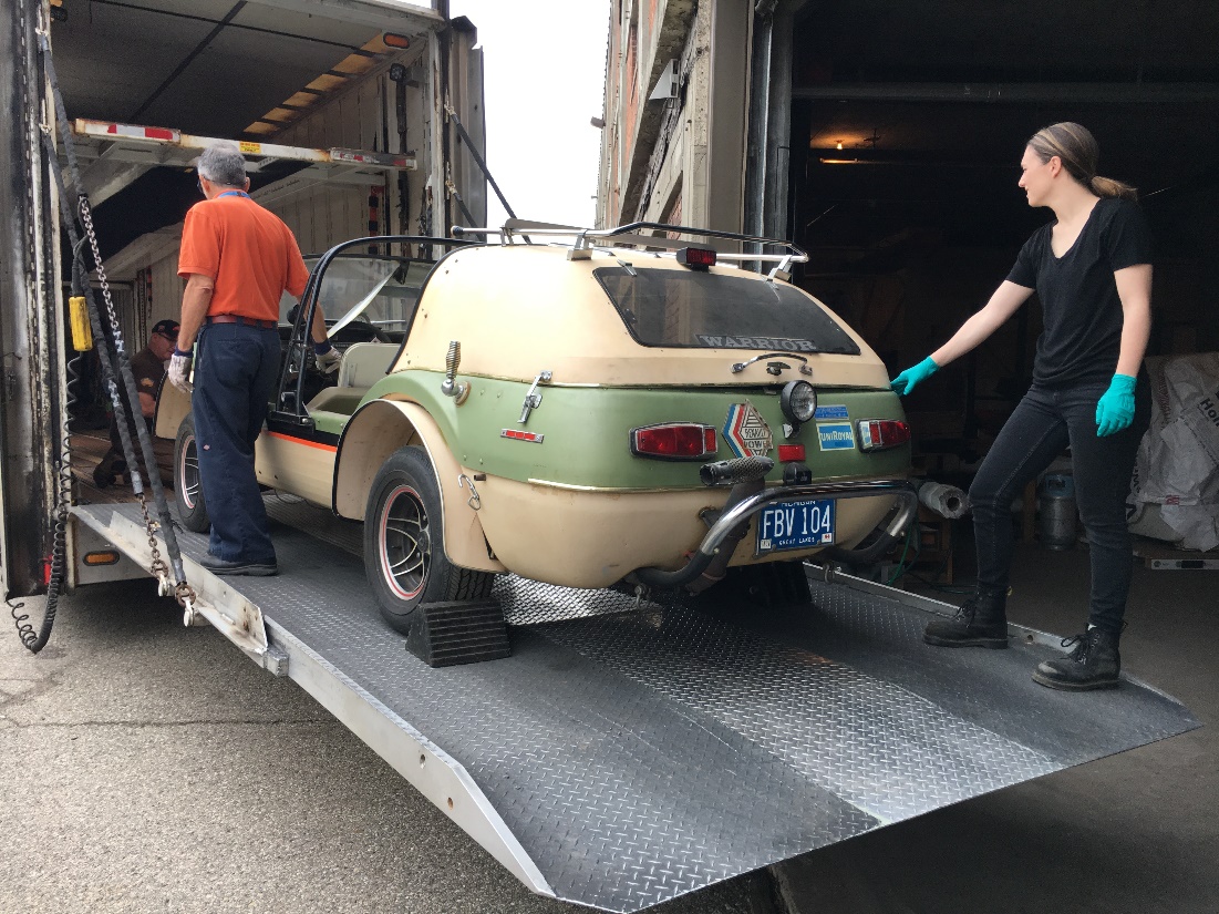 The Warrior is loaded into a semitruck Two people stand with an open beige-and-green car on the loading ramp of a truck; one kneels in front