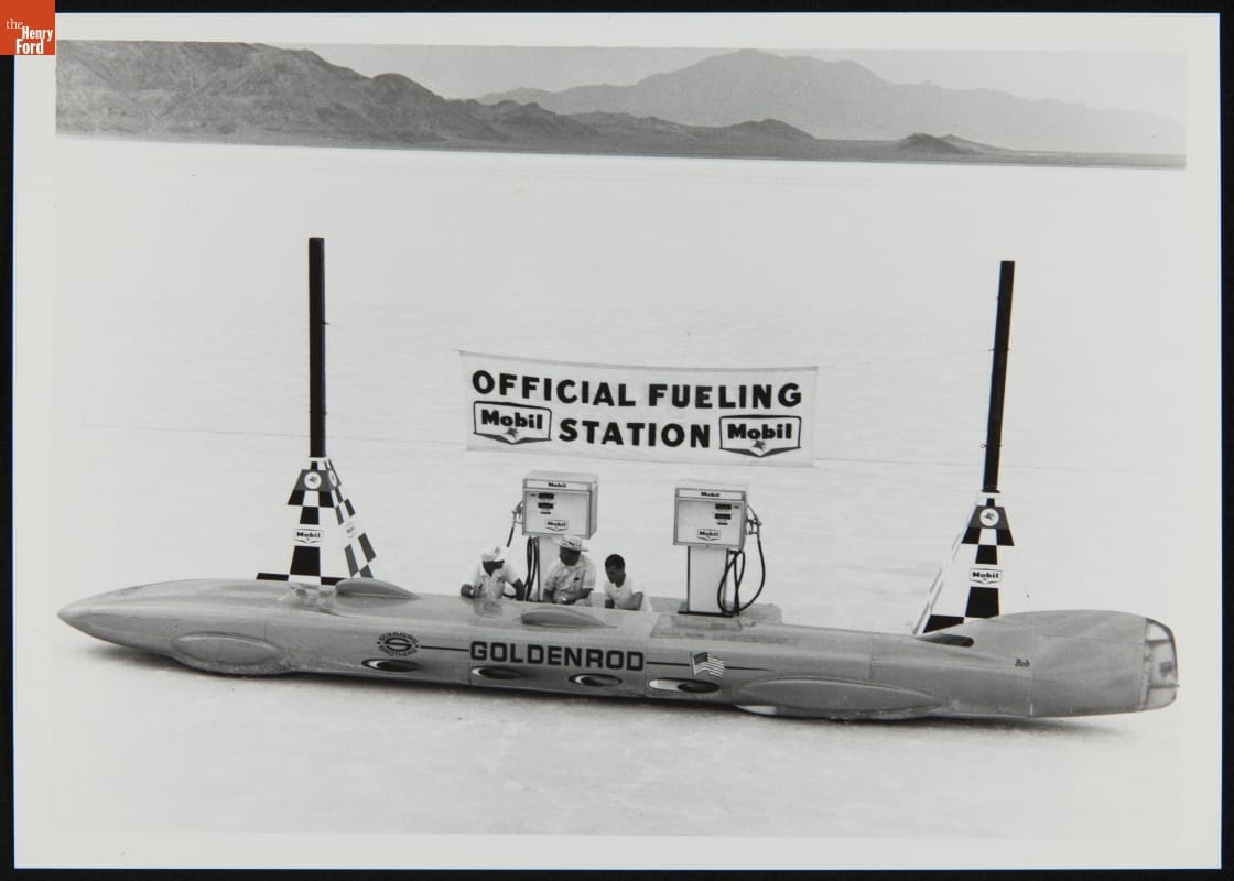 Long, low, race car at gas pumps under banner reading "Official Fueling Station," in open area with mountains in the background
