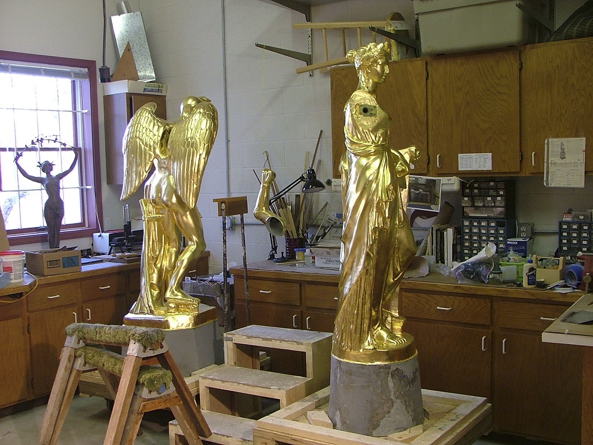 Newly gilded Father Time and the Muse. Two golden statues, one of a woman wearing flowing robes and one of a figure with wings, in a workroom