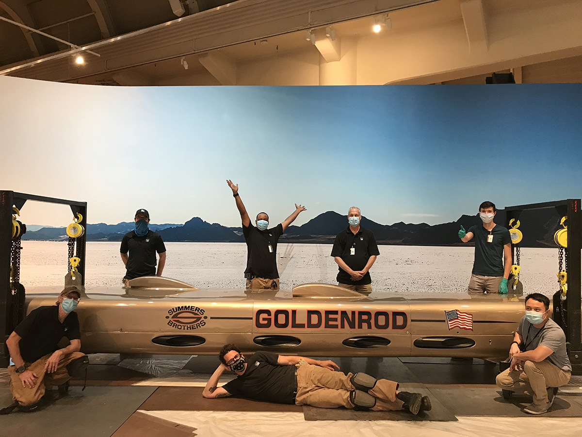 Exhibits and Conservation staff celebrate Goldenrod's final placement. Men wearing facemasks, one with arms raised triumphantly and one giving a thumbs-up, stand, kneel, and lie around a long, low, torpedo-shaped golden car in front of a backdrop of a salt flat, mountains, and blue sky