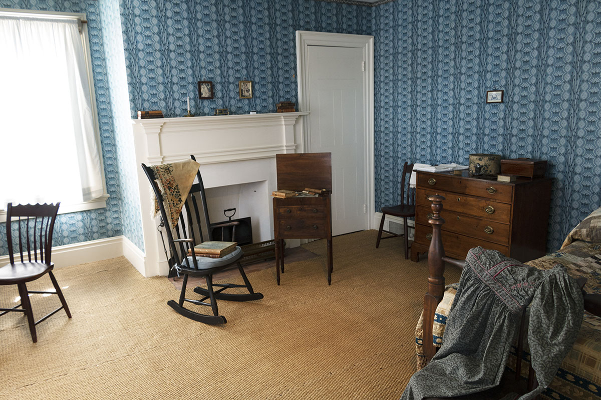 Room with patterned blue wallpaper containing fireplace, bed, chest of drawers, chairs