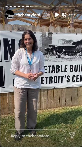 Woman in tan slacks and white shirt with lanyard around neck stands outside a wooden fence, behind which a building is partially erected