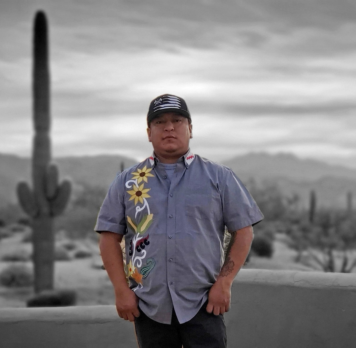 Chef Brian Yazzie Man in short-sleeved blue button-down shirt with embroidery of flowers and vegetables along one side stands in a desert landscape with cacti and mountains