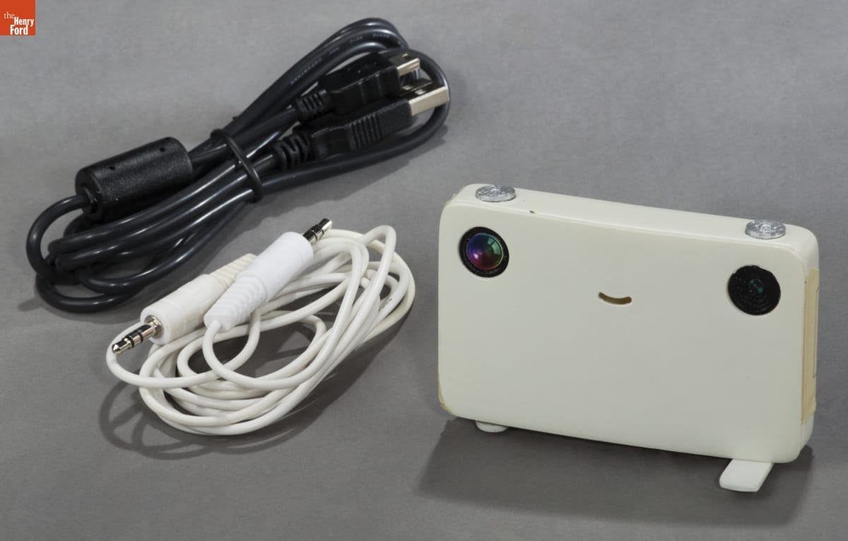 Model of "Pico - The Projector Camera," 2010 White rectangular device with a smiley face and two cords, one white and one black