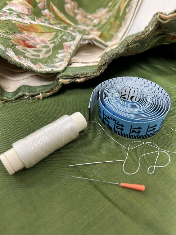 Small tools and thread are less likely to damage the delicate fabric A tape measure, spool of white thread, needle, and small auger lay on top of moss-green fabric with elaborate embroidery
