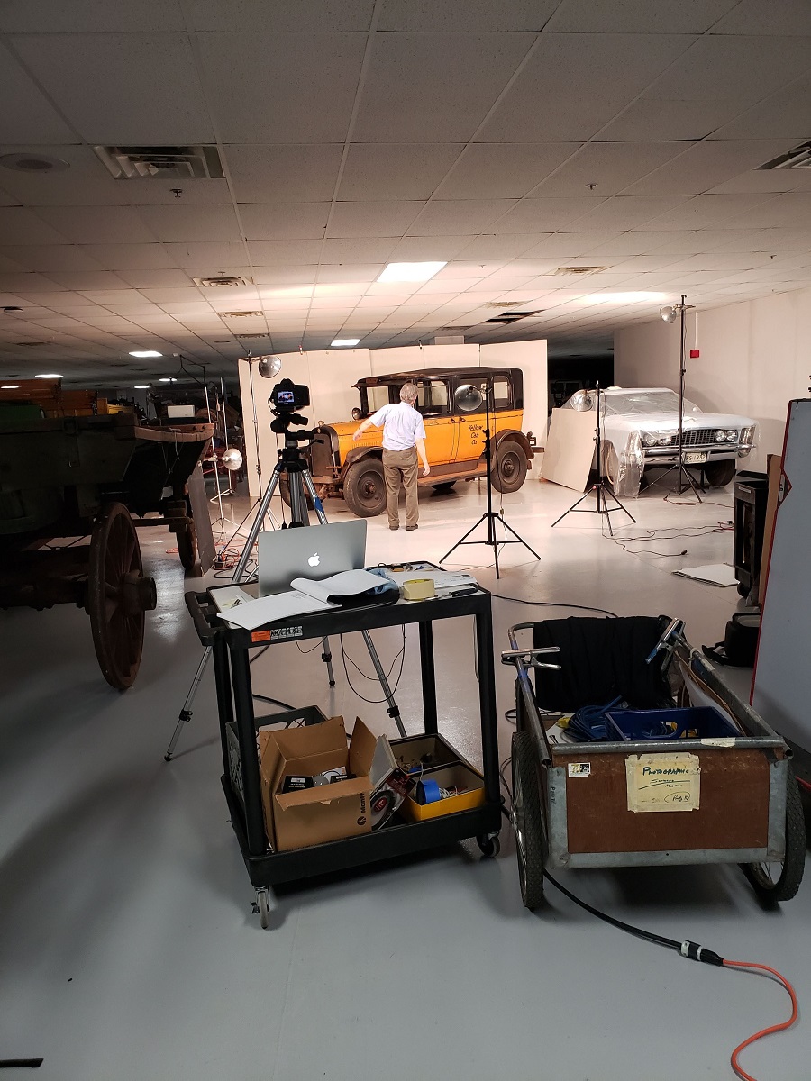Photographing the 1925 Yellow Cab taxicab A yellow car sits in a cramped space among other vehicles in front of a white background, with carts of photo equipment, a camera on a tripod, and lights on stands in the foreground