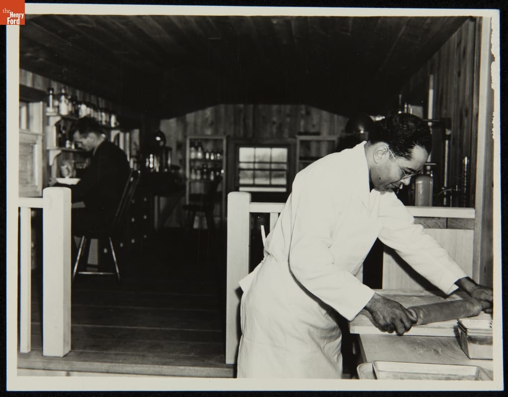 Man in white apron works with rolling pin while another man in background works with a mortar and pestle
