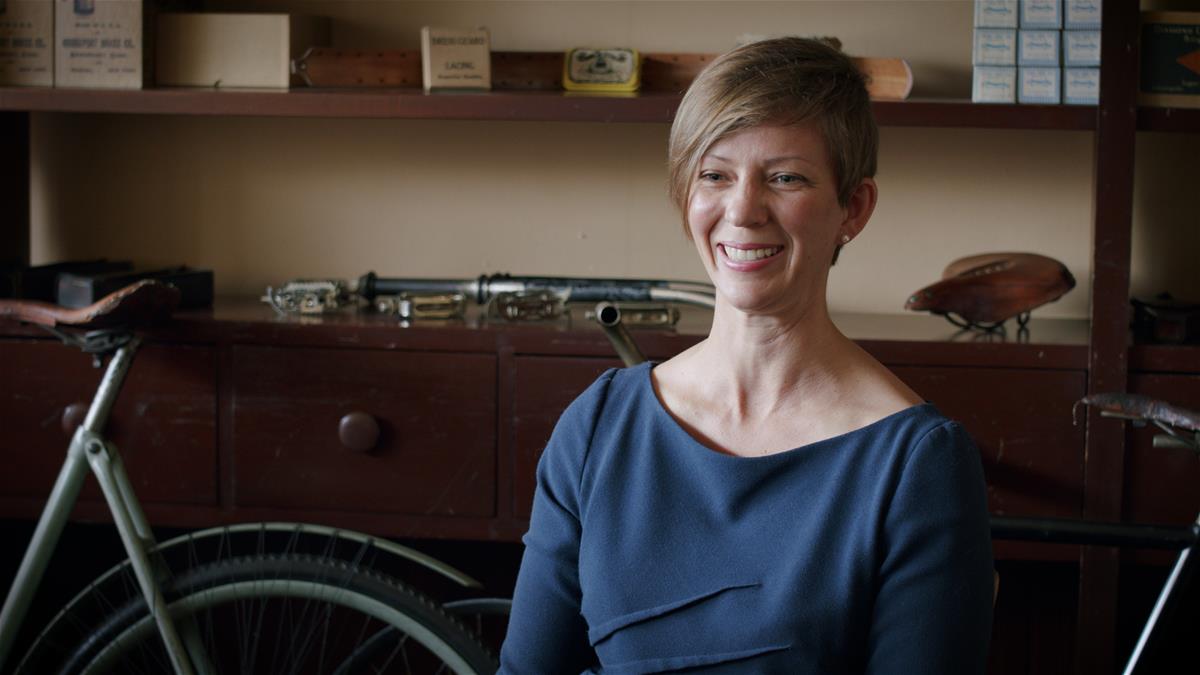 Jessica Robinson during an Interview for the Initiative for Entrepreneurship, 2020 Woman in blue top smiling, bicycle and counter/shelves partially visible behind her