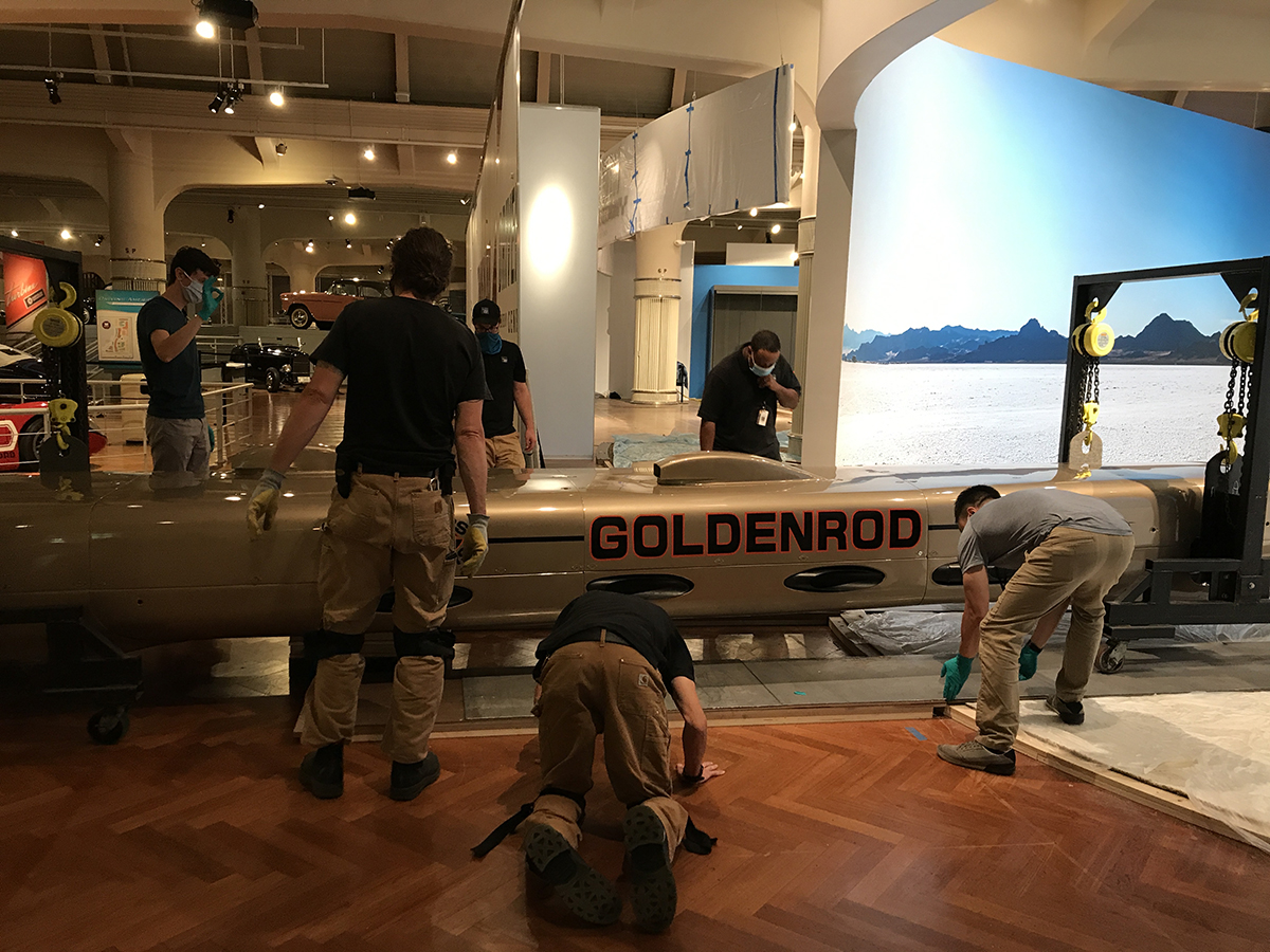 Halfway there! Group of men wearing facemasks stand and kneel around a long, low, torpedo-shaped golden car