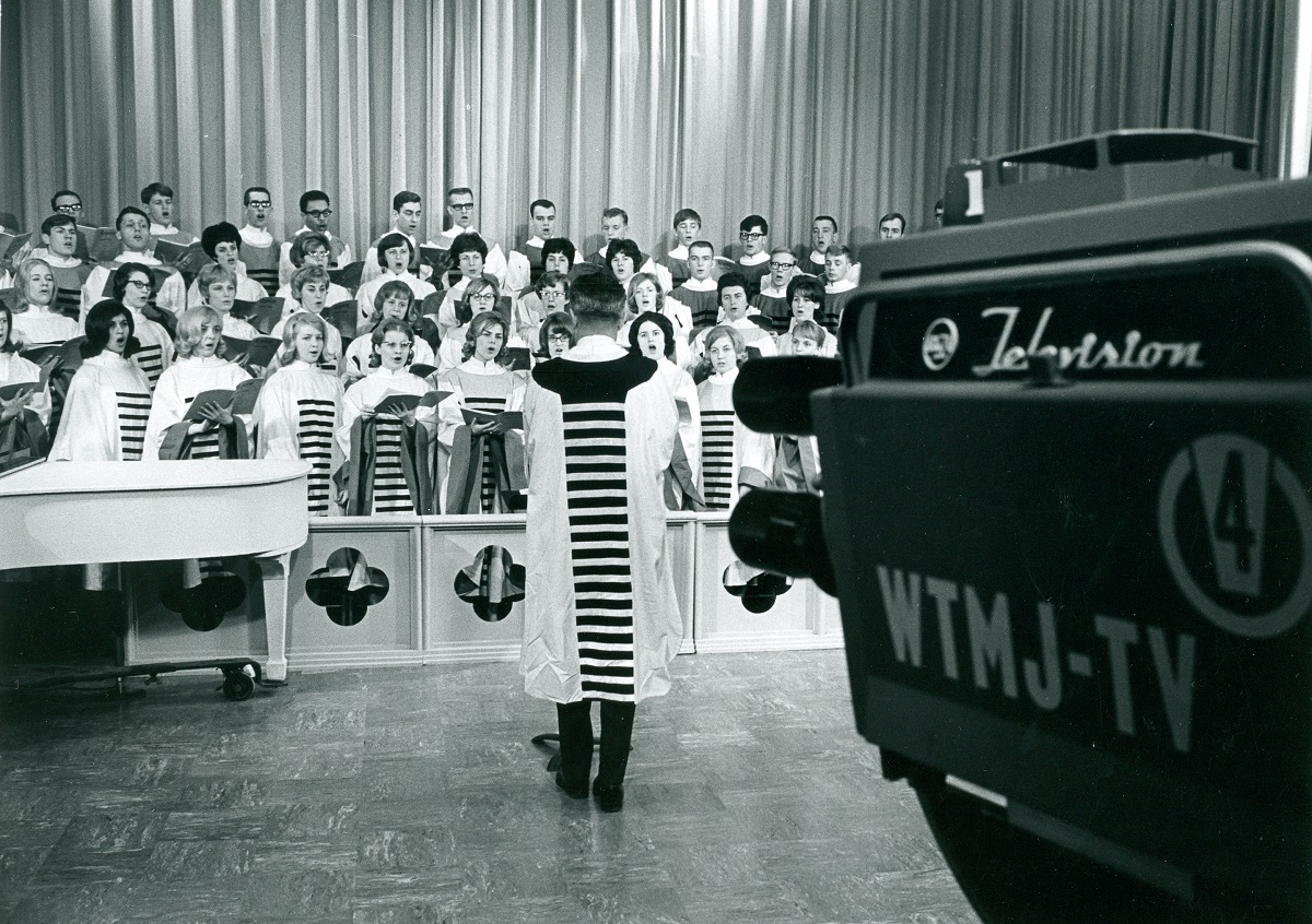 Hope College Chapel Choir Recording at Milwaukee’s WTMJ-TV Black-and-white photo with TV camera in foreground, pointing at group of people in robes being conducted by another person in a robe