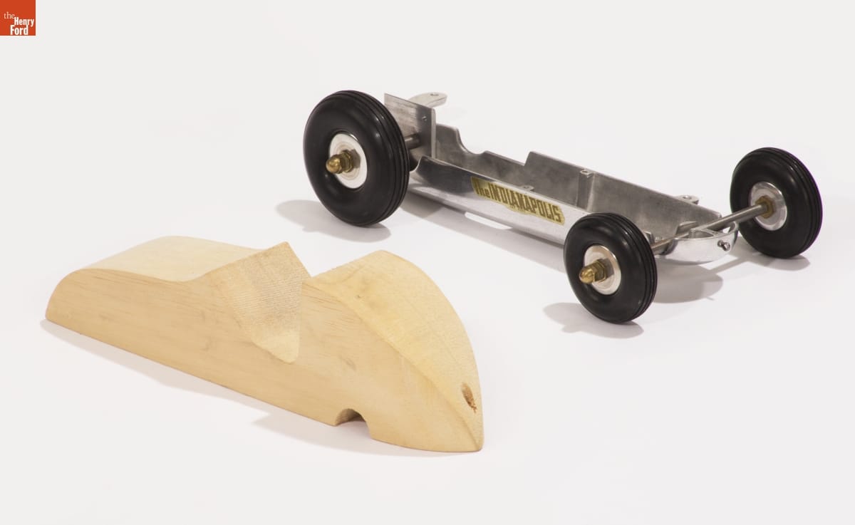 Korn "Indianapolis 29" Gas-Powered Racing Tether Car Kit, 1948 Metal model car chassis sitting next to carved, unpainted block of wood in the shape of a car body