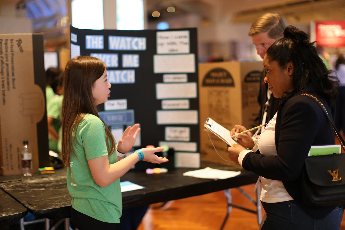 A student pitches their invention to volunteer judges A young girl in a green t-shirt talks to two other people in front of a trifold display board on a table