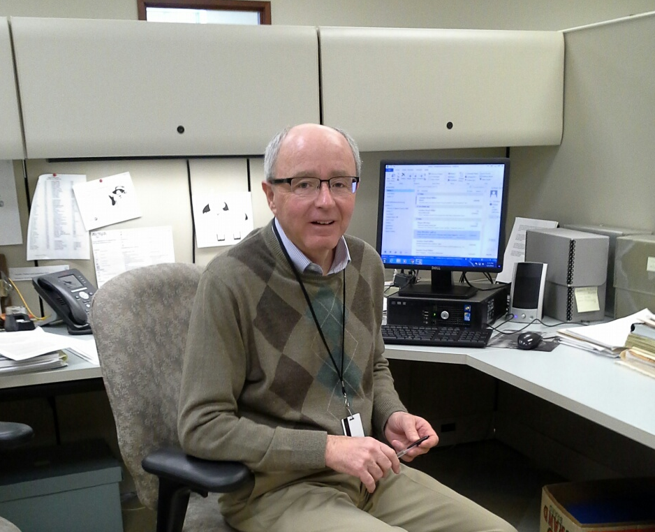 Man with glasses and white hair wearing an an argyle sweater and khaki pants sits at an office cubicle with a computer and silver archival boxes nearby