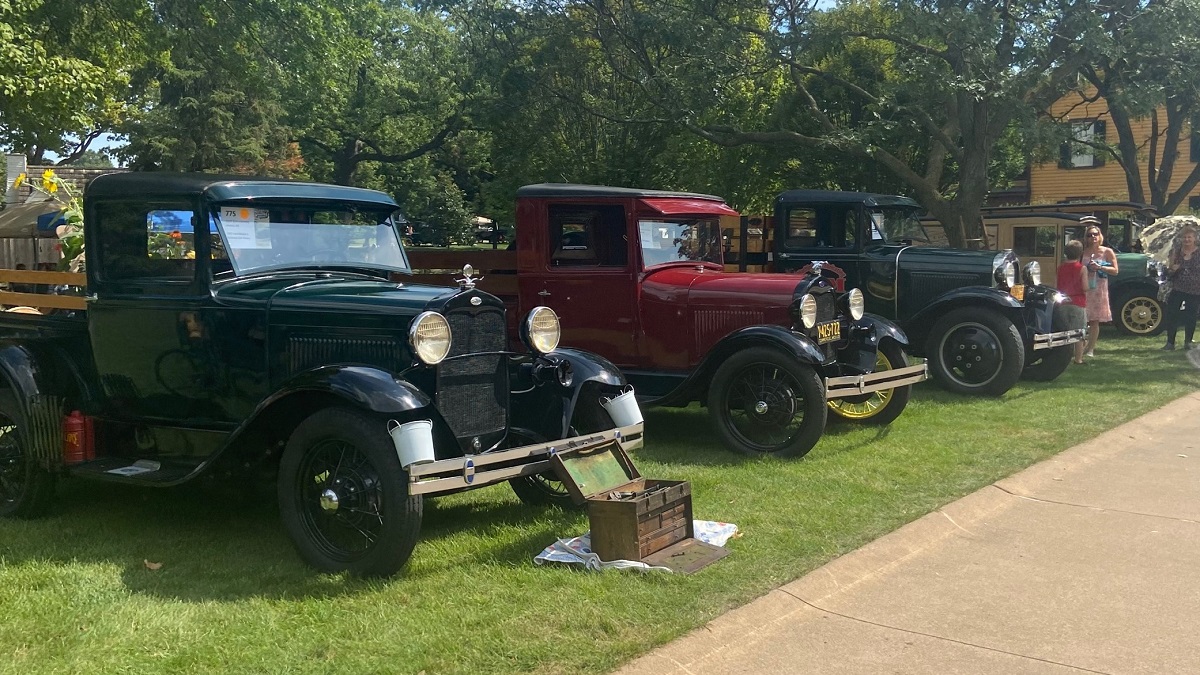 Commercial vehicles line Christie Street in Greenfield Village during Old Car Festival 2022 Old-fashioned trucks are parked in a line on a green lawn with large trees and buildings behind them and people nearby
