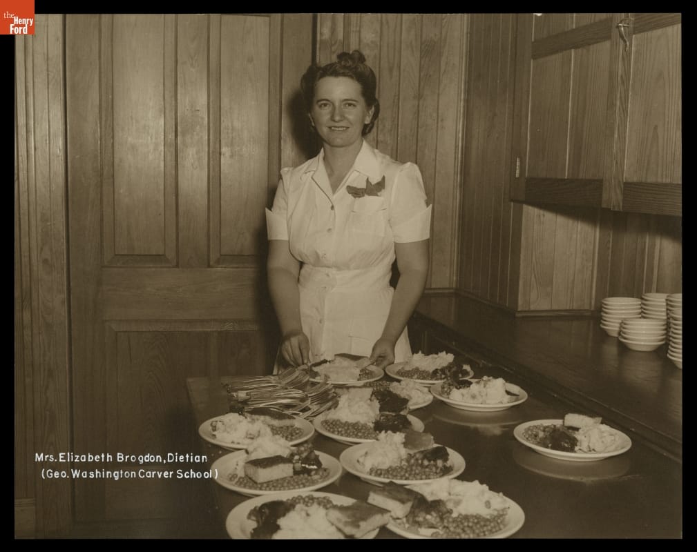 White woman stands behind table laden with plates of food in wood-paneled room