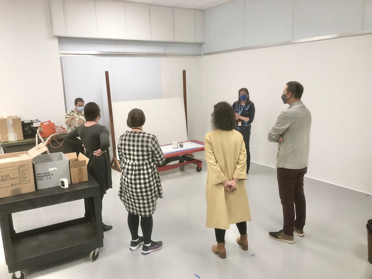 Group of people wearing masks stand in a circle in the corner of a room with several carts filled with boxes nearby
