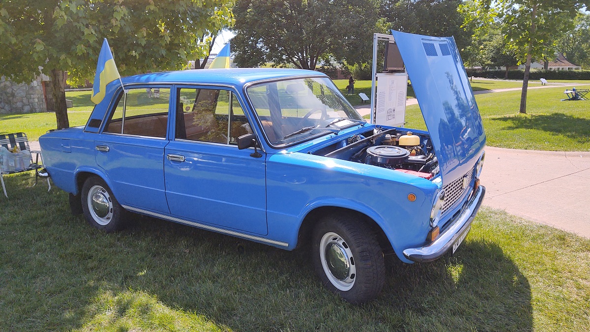 1978 VAZ 21011 sedan built in the Soviet Union at Motor Muster 2022 Small, boxy, sky-blue car with hood open on a lawn among trees and walkways