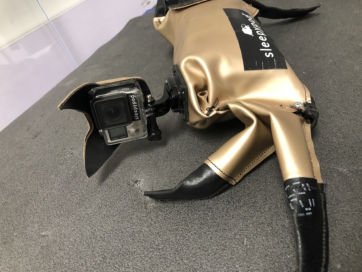 Sleepypod "CLEO" Crash-Test Cat, lying on side Champagne-colored fabric or plastic cat figure, with text and circular shape on side and camera where face would be, lying on side