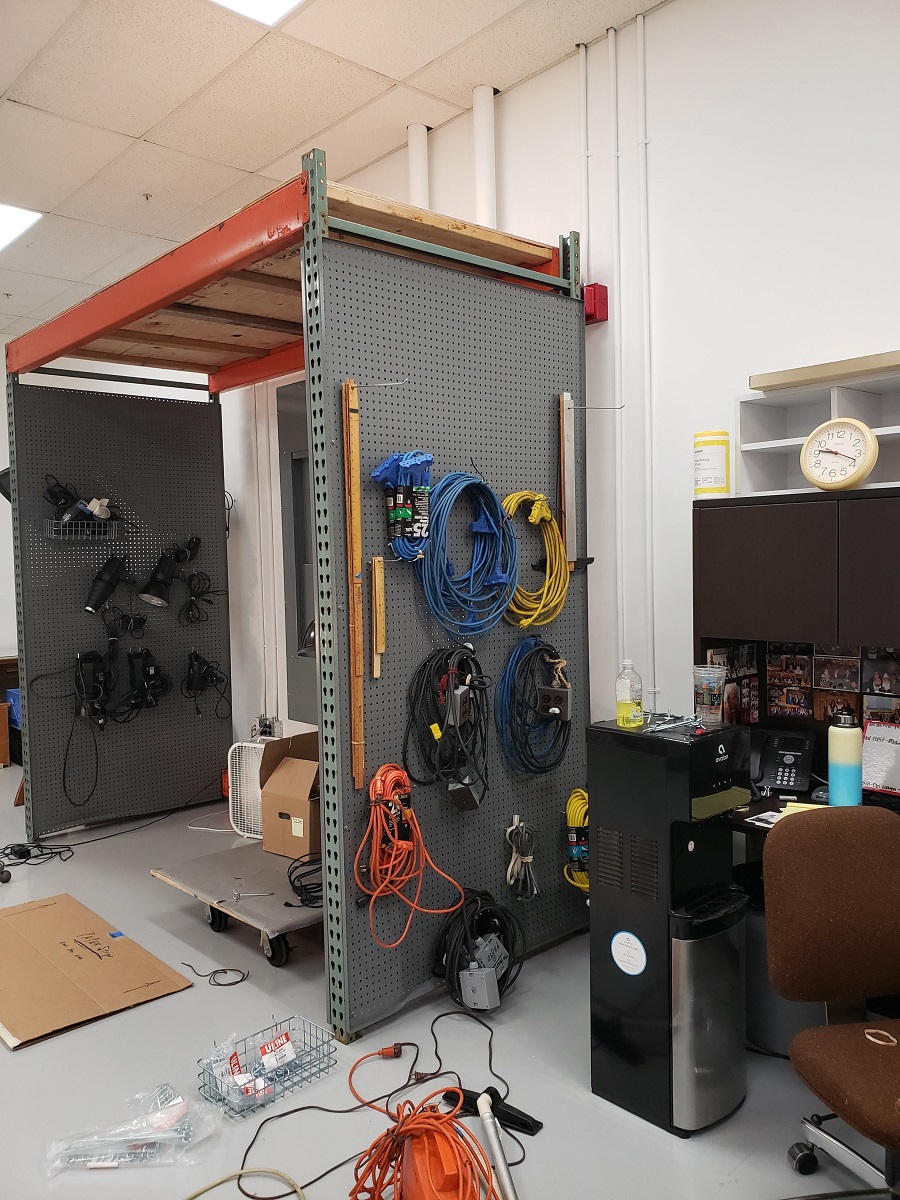 Cluttered room with pallet racking covered in pegboard with cables and wires hanging on it