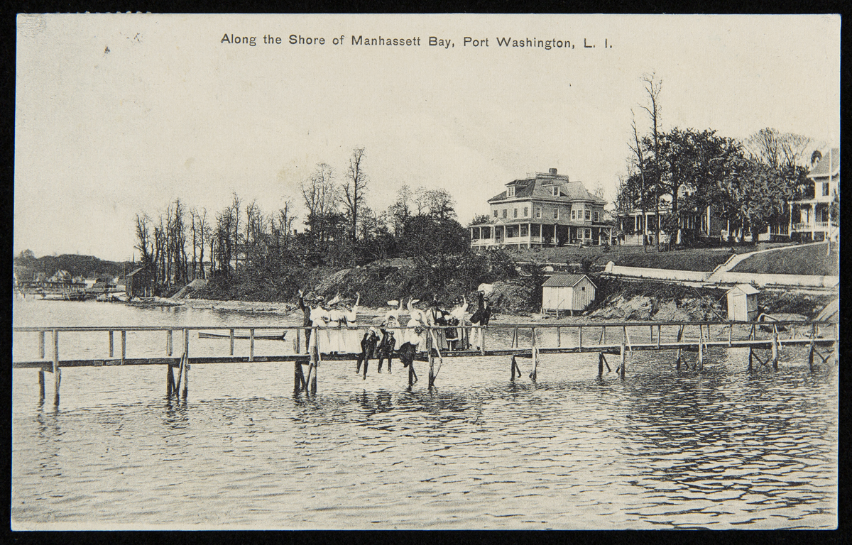 Along the Shore of Manhassett Bay, Port Washington, L.I., October 4, 1909 People wave from long, wooden dock over a body of water with houses visible along shoreline behind them; also contains text
