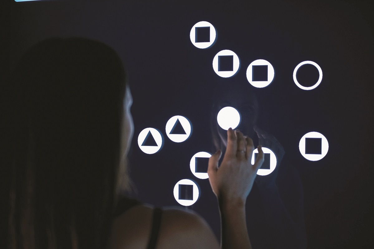 Sensory station Woman stands with hand up to a black digital board with lighted circles, triangles, and squares within white circles