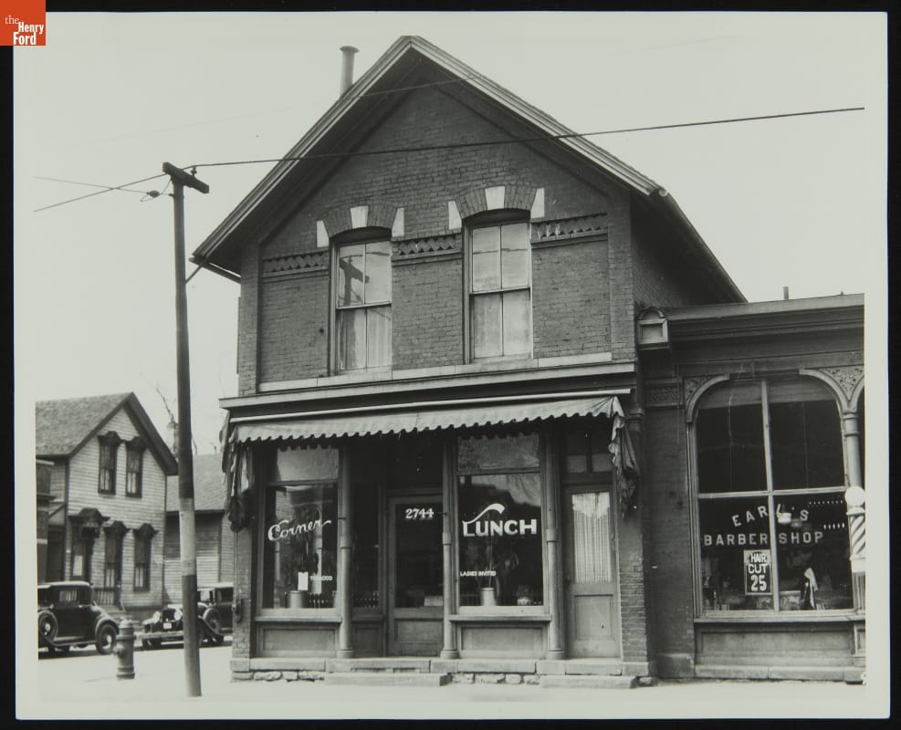 Black-and-white photo of street scene, focused on two-story brick building with business windows on first floor