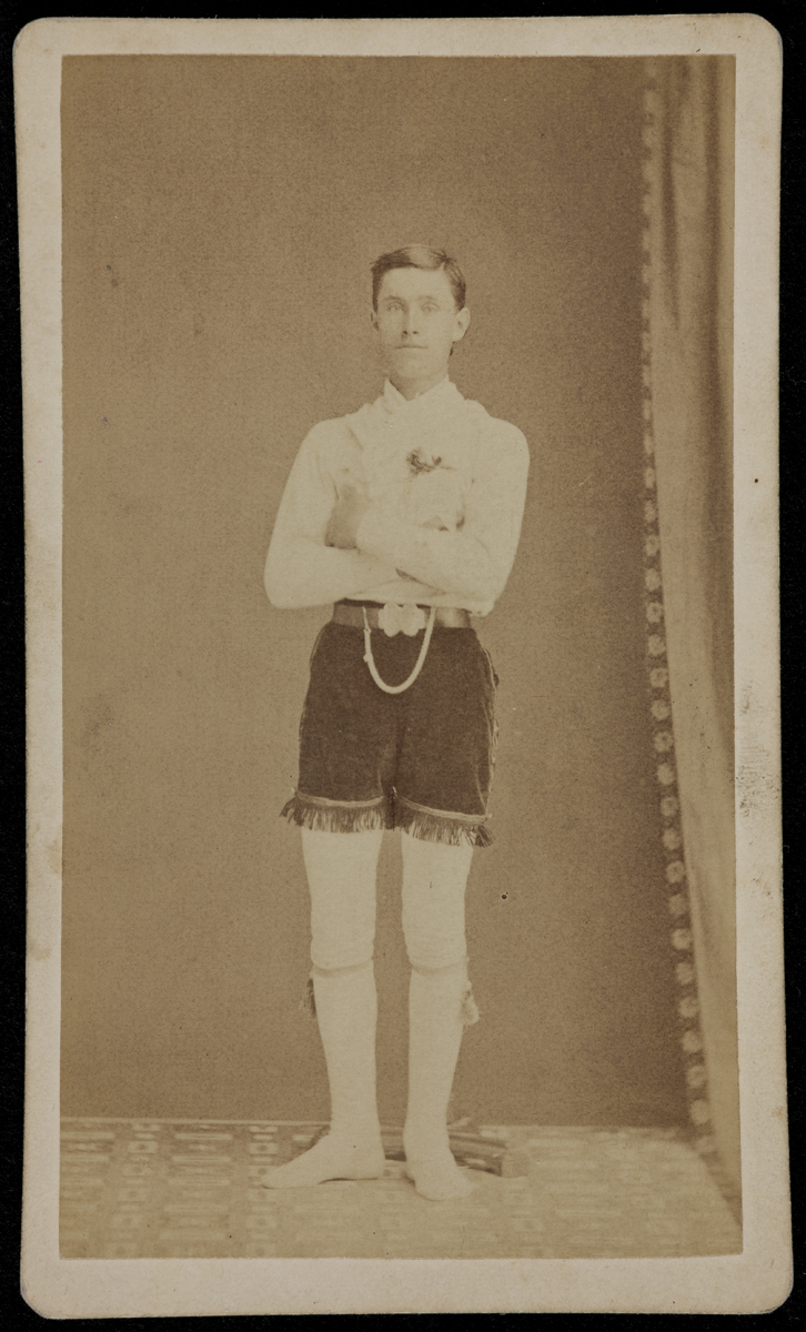 Black-and-white photo of man standing with arms crossed, wearing white long-sleeved shirt, white tights (?), and dark velvet shorts with fringe