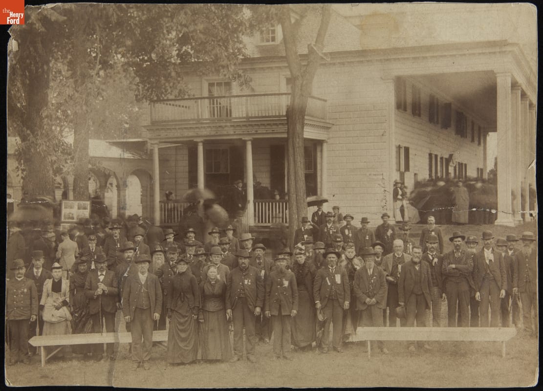 Sepia-toned photo of a large group of men and women in 19th-century clothing standing in front of a two-story wooden house with large porch and columns