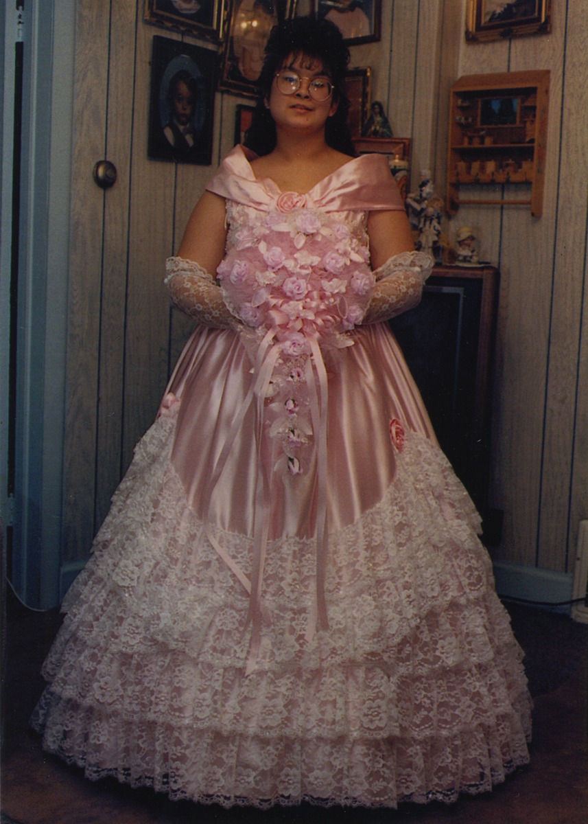 Maritza Garza in Her Quinceanera Dress, 1992 Woman with long dark hair and glasses wearing pink gown with tiers and white lace holds an elaborate flower arrangement