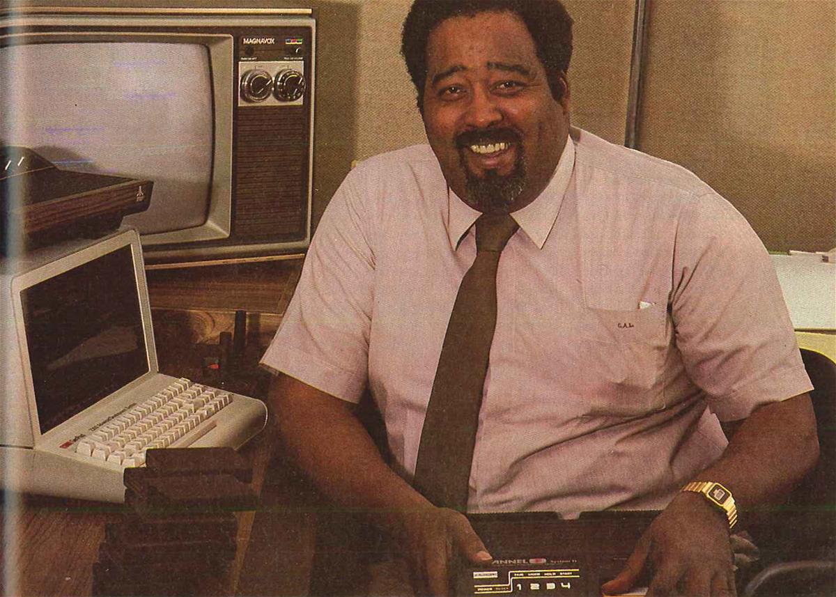 Jerry Lawson, circa 1980 Black man with beard and mustache in shirt and tie sits by a computer and television and smiles at camera