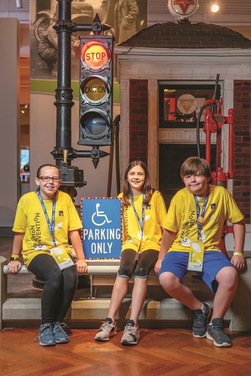 Team Scandicapped, Invention Convention U.S. Nationals President's Choice award winners Three children in yellow t-shirts sit in front of a old-fashioned traffic light and small brick gatehouse in a larger building; one holds a blue and white "handicapped" parking sign