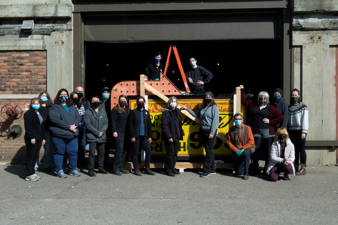 Team photograph with the last artifact to leave the warehouse A group of people wearing masks pose for a photograph in front of, and behind, on a ladder, a large sign