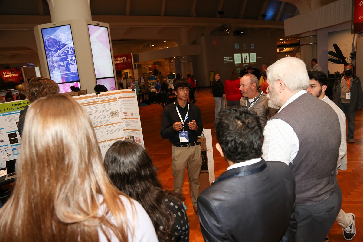 A student pitches their invention to volunteer judges A young man stands by a trifold display board, talking to a group of people circled around him