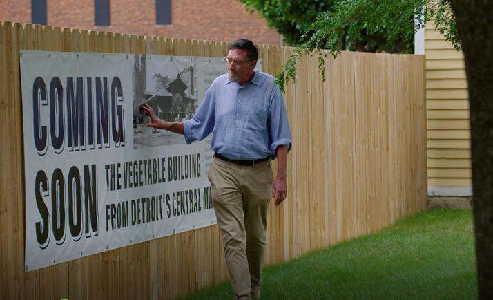 Man in blue button-down shirt and khaki pants walks along wooden fence with large white banner reading 